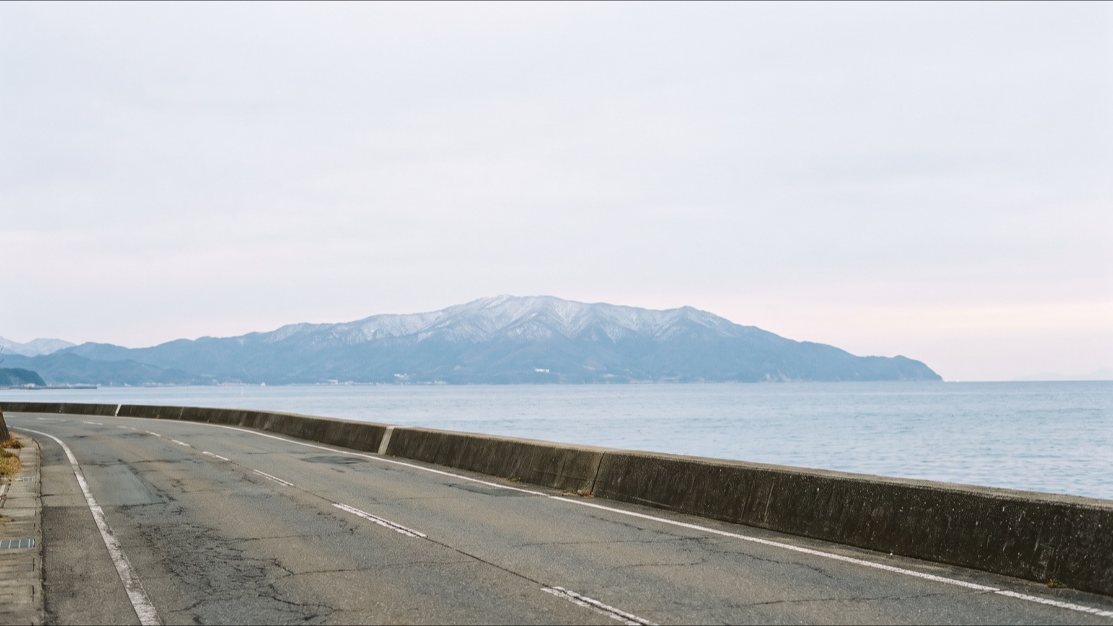 日本海沿いの人のいない道と防波堤。遠くに雪をかぶった山並みが見える
